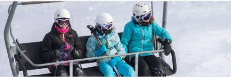 Three young skiers, wearing helmets and winter gear, sit on a ski lift, ready for an adventure on the slopes. Snow-covered landscape surrounds them.