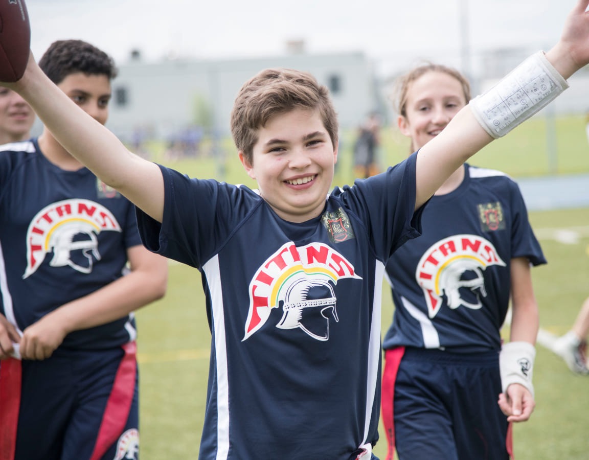 A smiling boy in a Titans football uniform celebrates while holding a football, with teammates in the background on a grassy field.