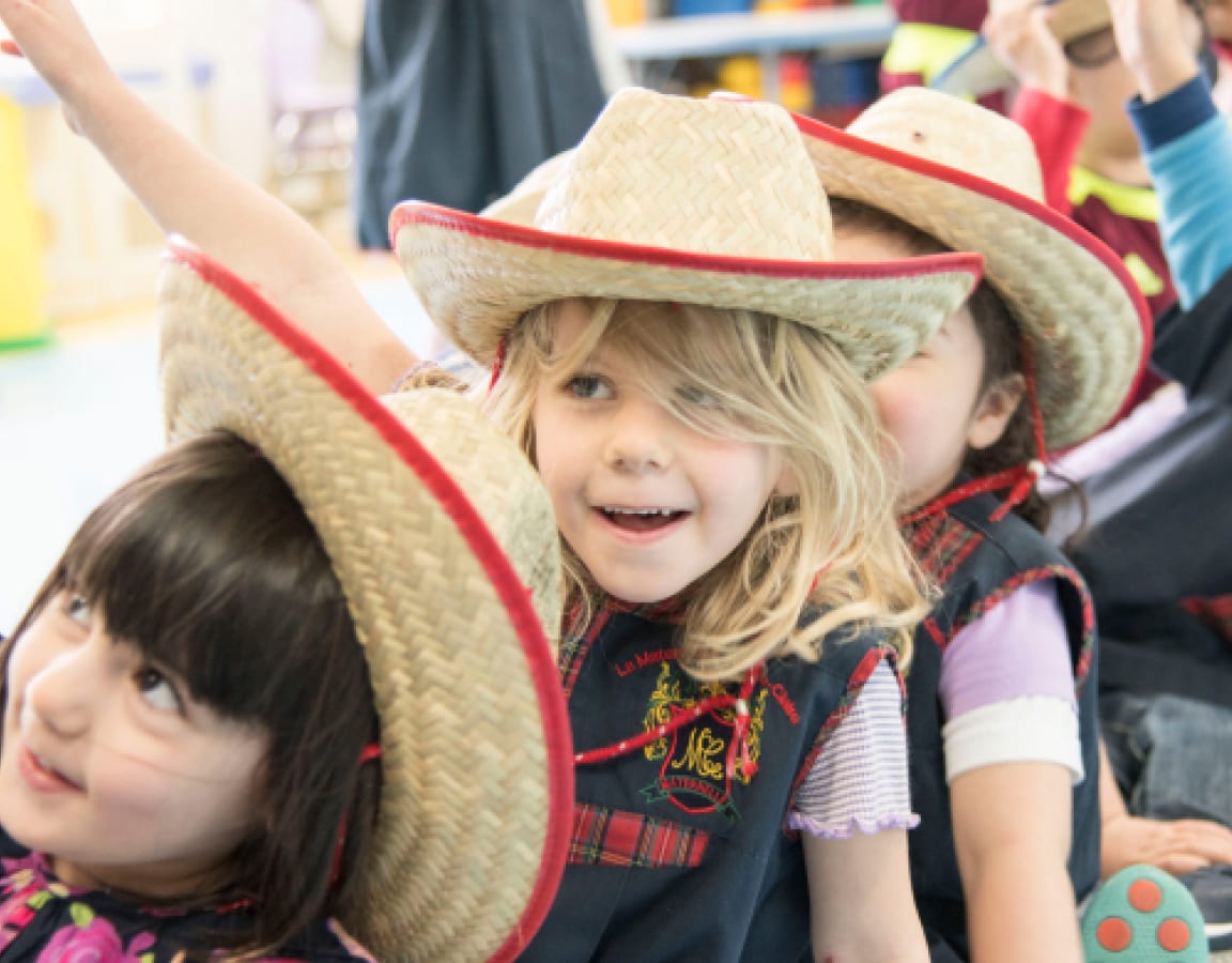 Children in a classroom wear straw hats, smiling and engaged in an activity, showcasing a fun and lively learning environment.