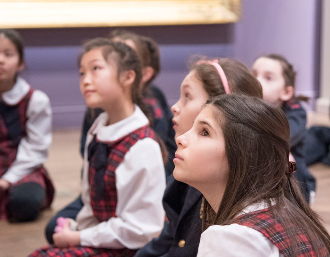 School children in plaid uniforms sit attentively on the floor of an art museum, gazing up at an unseen exhibit, embodying curiosity and engagement.