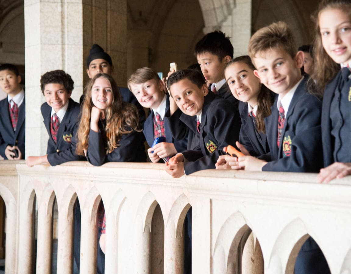 A group of smiling students in formal attire leans against a stone railing, posing for a photo in a grand building, reflecting a lively school outing.