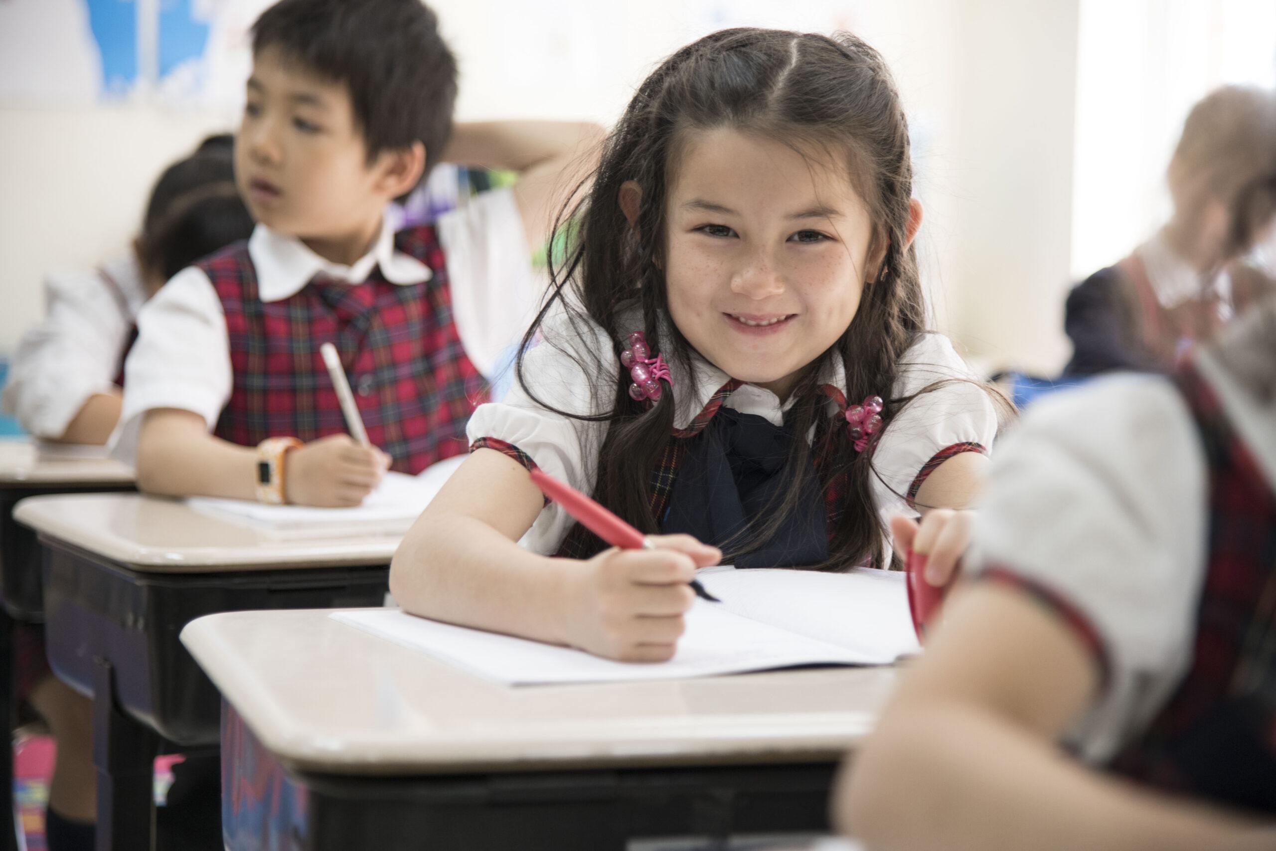A young girl in a school uniform smiles while writing in a notebook at her desk. Other students are engaged in the background, highlighting a classroom setting.