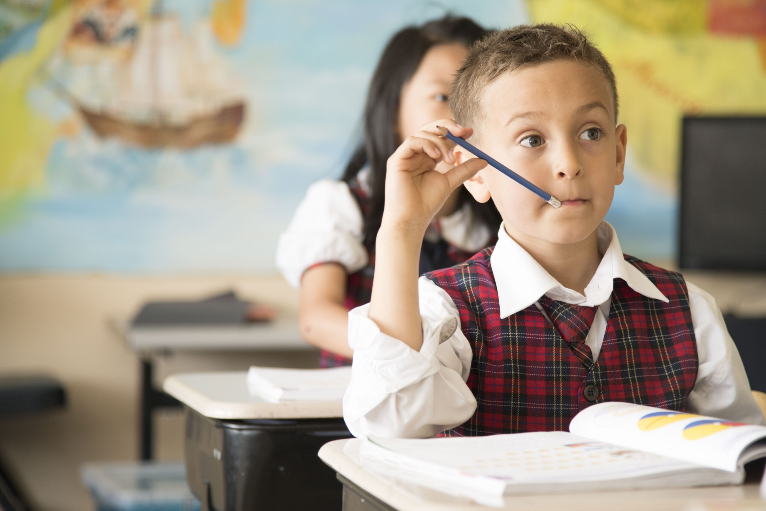 A student in a plaid vest thoughtfully holds a pencil to his mouth while seated at a desk, surrounded by classroom materials.