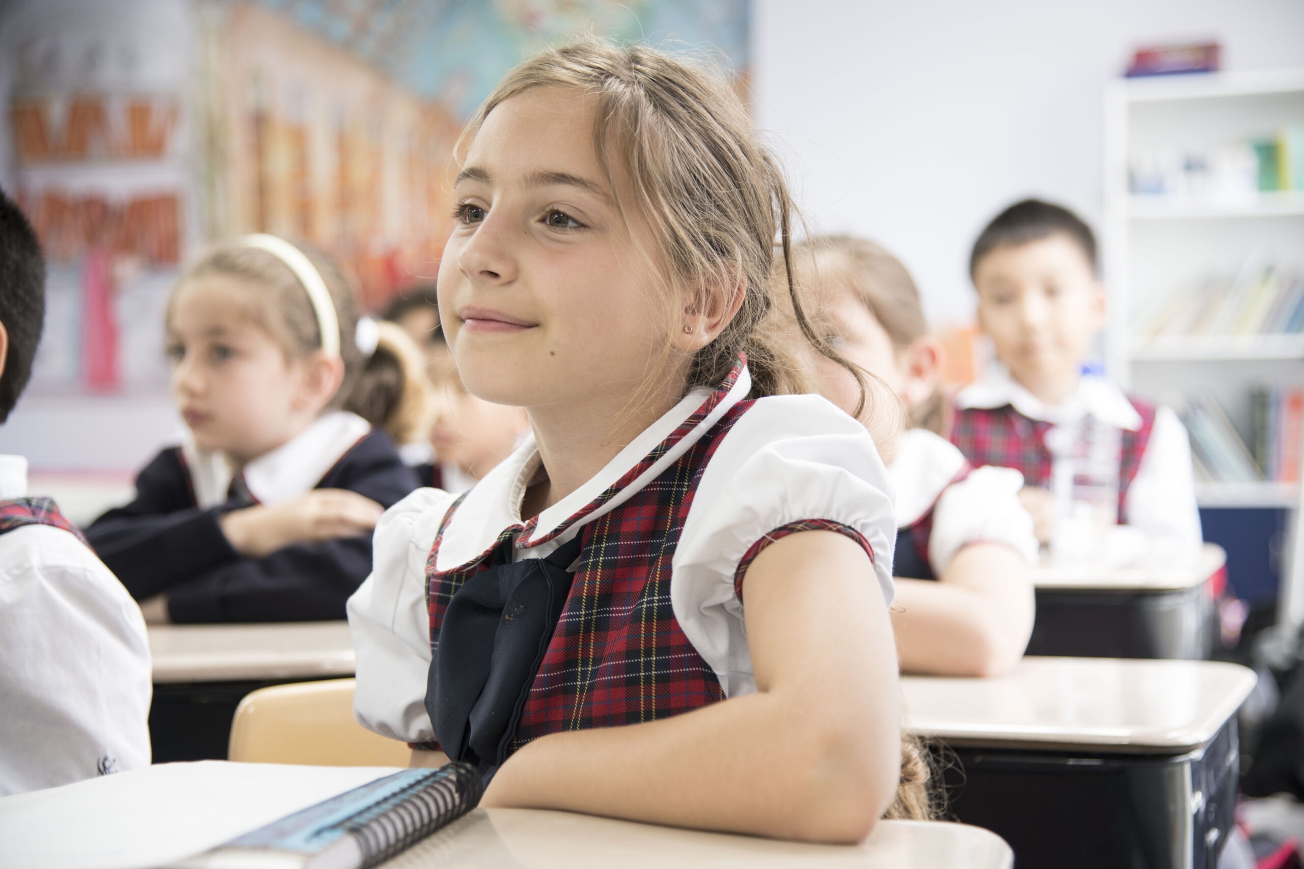 A school girl, wearing a plaid uniform, sits attentively at her desk in a classroom, surrounded by classmates engaged in learning.