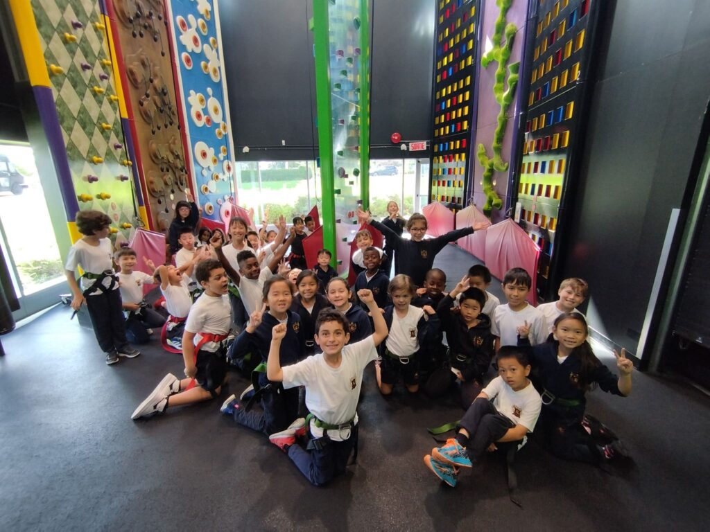 A diverse group of children poses excitedly in a climbing gym, showcasing vibrant climbing walls and harnesses. The image captures their enthusiasm for the activity.