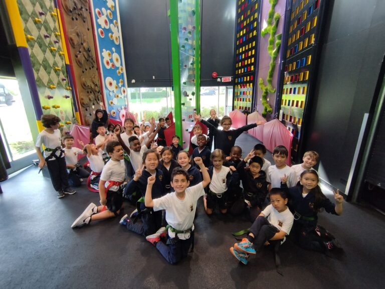 A diverse group of children poses excitedly in a climbing gym, showcasing vibrant climbing walls and harnesses. The image captures their enthusiasm for the activity.