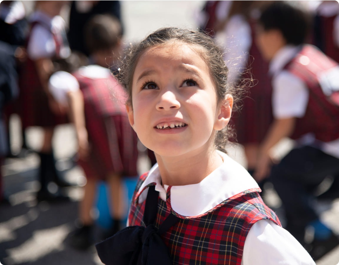 A young girl in a plaid school uniform looks upward with a curious expression, surrounded by classmates outdoors on a sunny day.