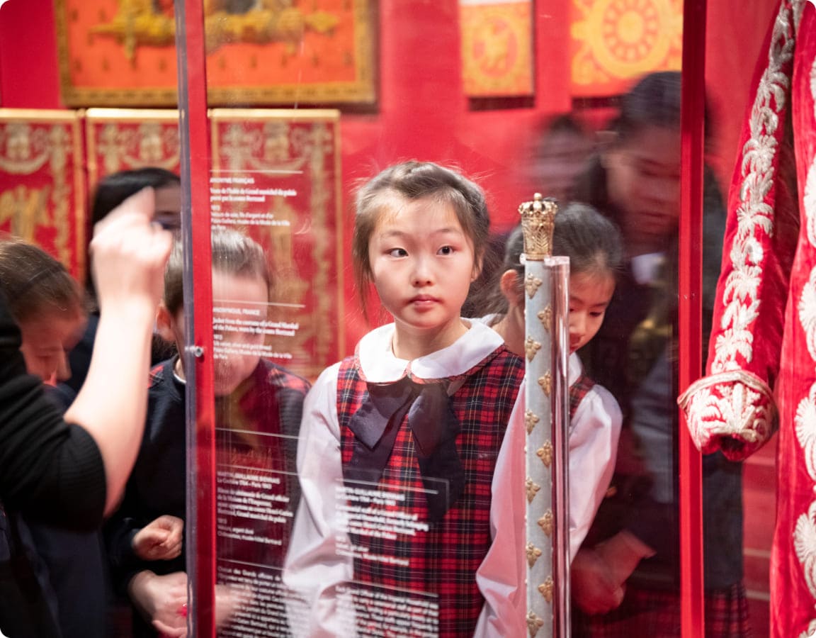 A young girl in a plaid outfit observes an exhibit in a museum, surrounded by peers and a glass display showcasing historical garments.