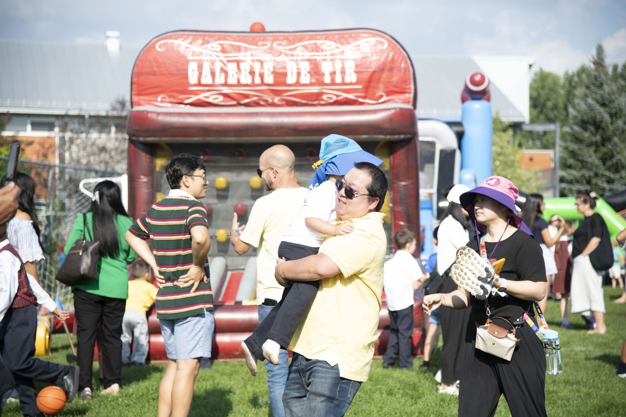 A father carries his child in a crowded park during a festive event, with a bouncy castle and various activities in the background.