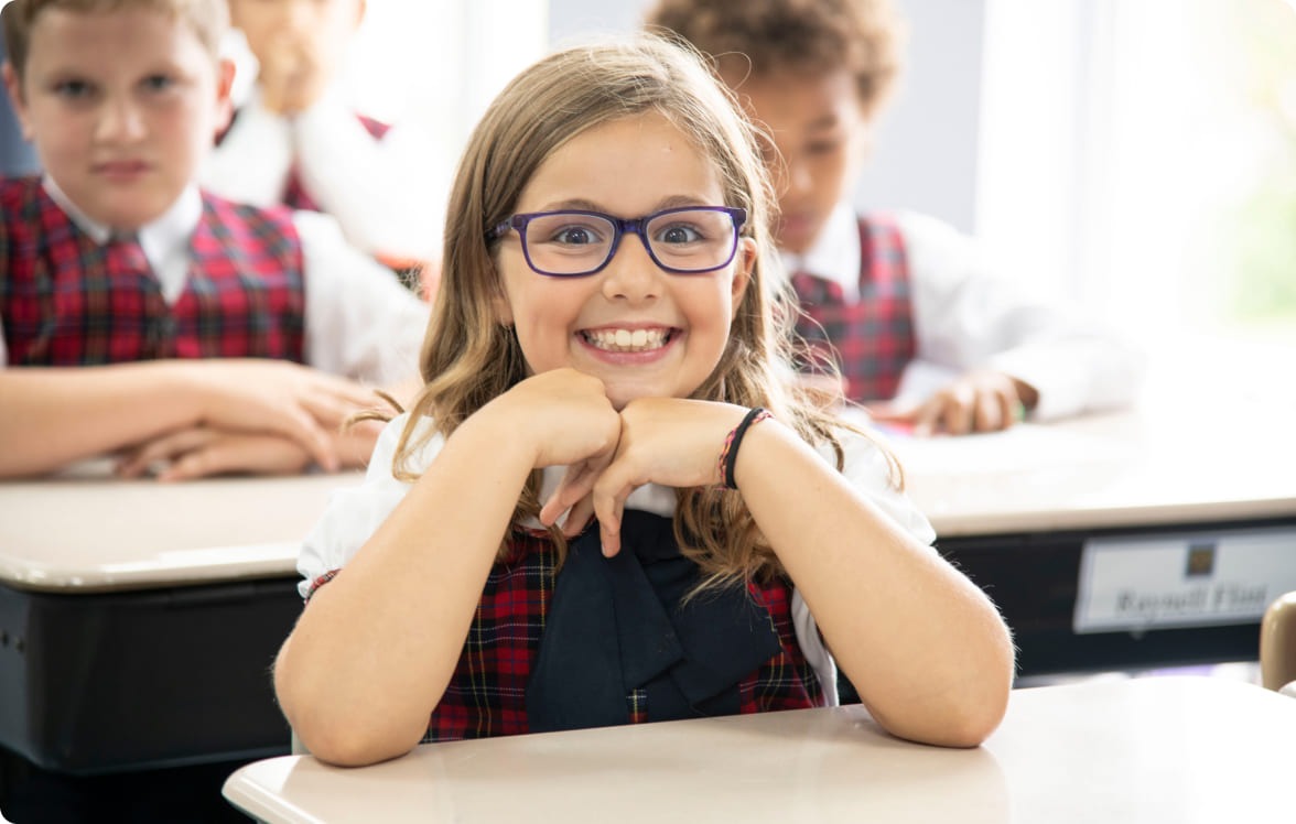 Smiling girl wearing glasses and a school uniform sits at a desk in a classroom, illustrating a positive learning environment.