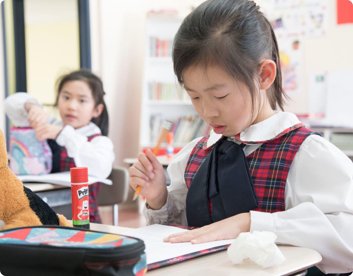 A young girl in a plaid school uniform focuses on drawing at her desk, while another girl in the background adjusts her backpack.