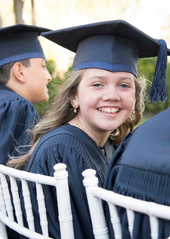 Smiling girl in a graduation gown and cap. She sits among fellow graduates, embodying joy and pride during the ceremony.