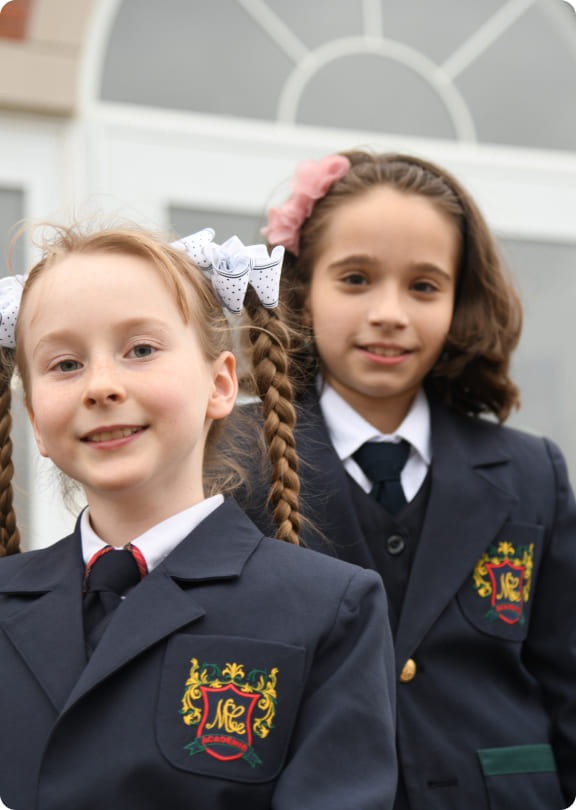 Two girls in formal school uniforms pose together, smiling. The girl in front has braided hair with a bow; the other has wavy hair.