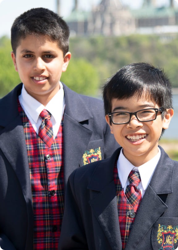 Two boys in formal attire stand outdoors, smiling against a blurred background. They wear blazers over plaid vests and ties, exuding a sense of camaraderie.
