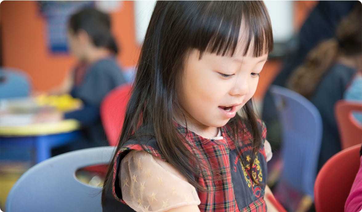 A young girl in a plaid dress smiles while engaged in an activity at a colorful classroom, reflecting joy and curiosity in a learning environment.
