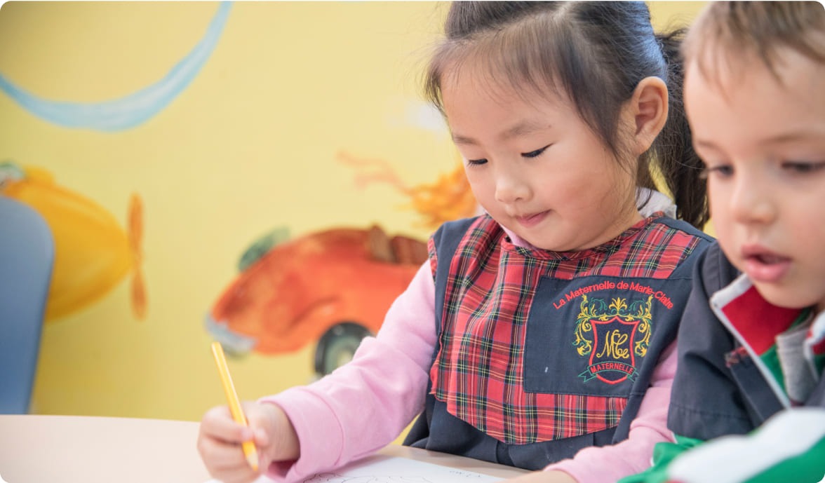 A young girl in a plaid smock colors with a pencil at a table, while a boy sits nearby, focused on his activity. A colorful mural adorns the background.