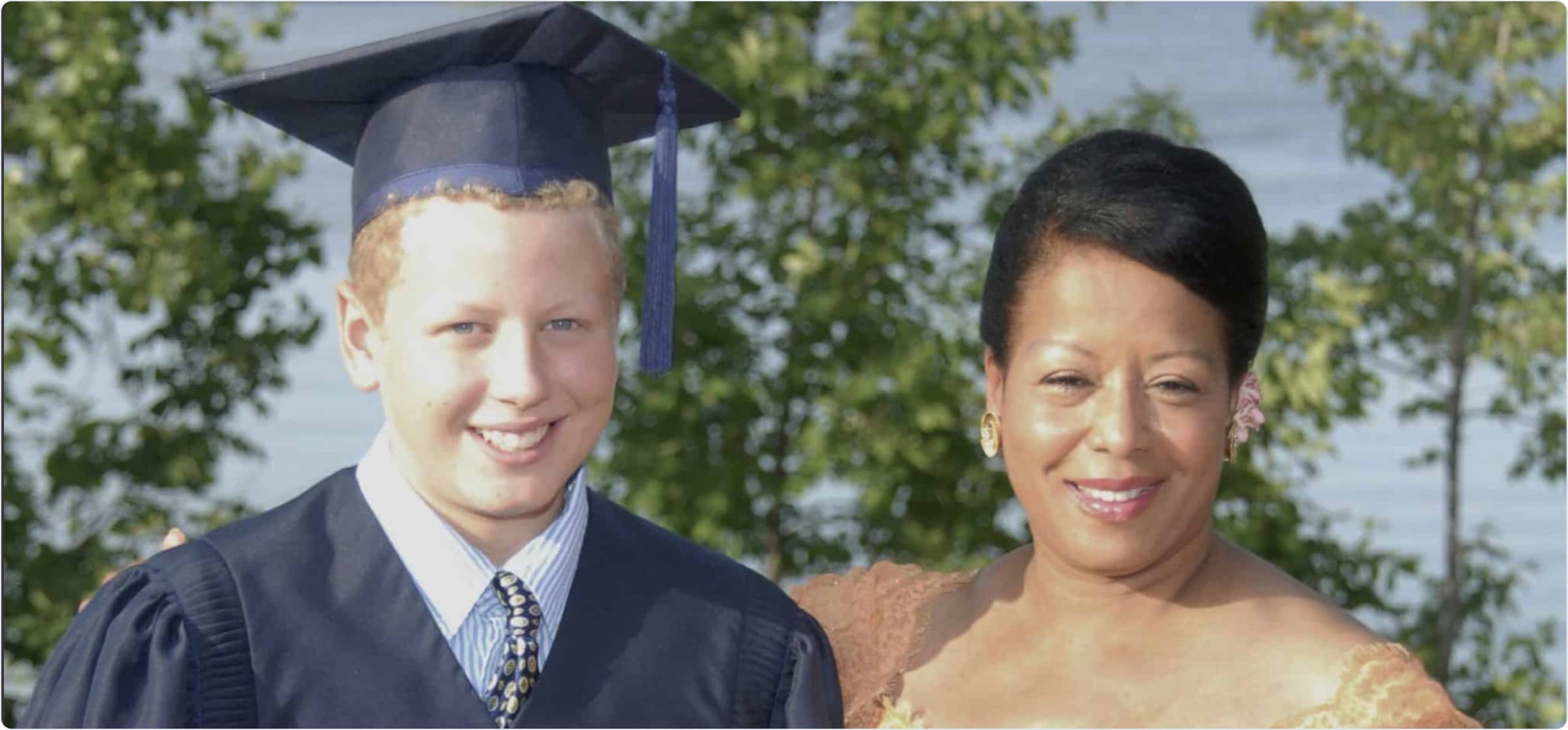 A smiling young graduate in a cap and gown stands beside a woman dressed in formal attire, both posing outdoors near trees.