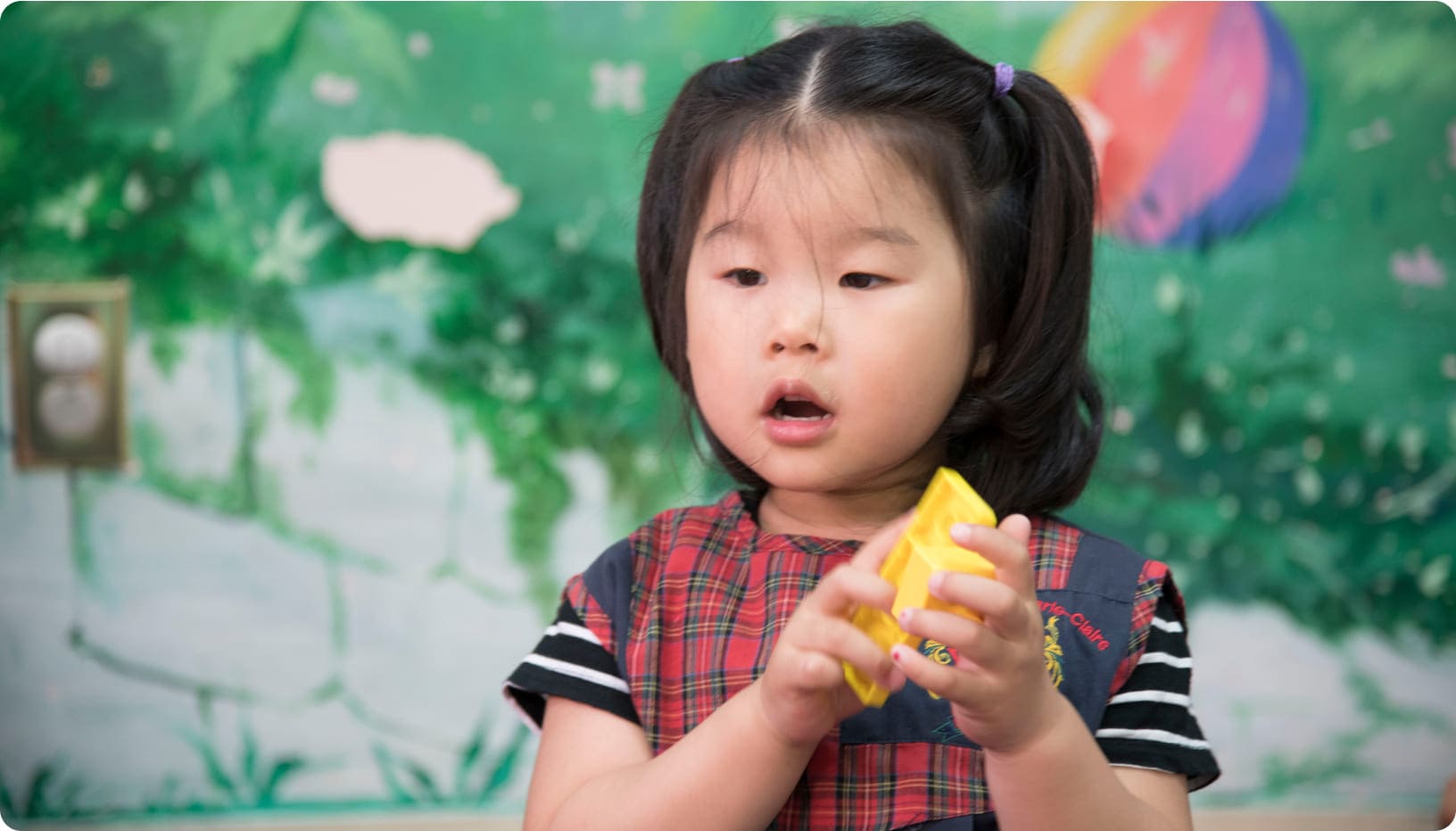 Young girl with pigtails holds a yellow toy, focused and engaged, against a colorful mural backdrop. The image depicts playful learning.