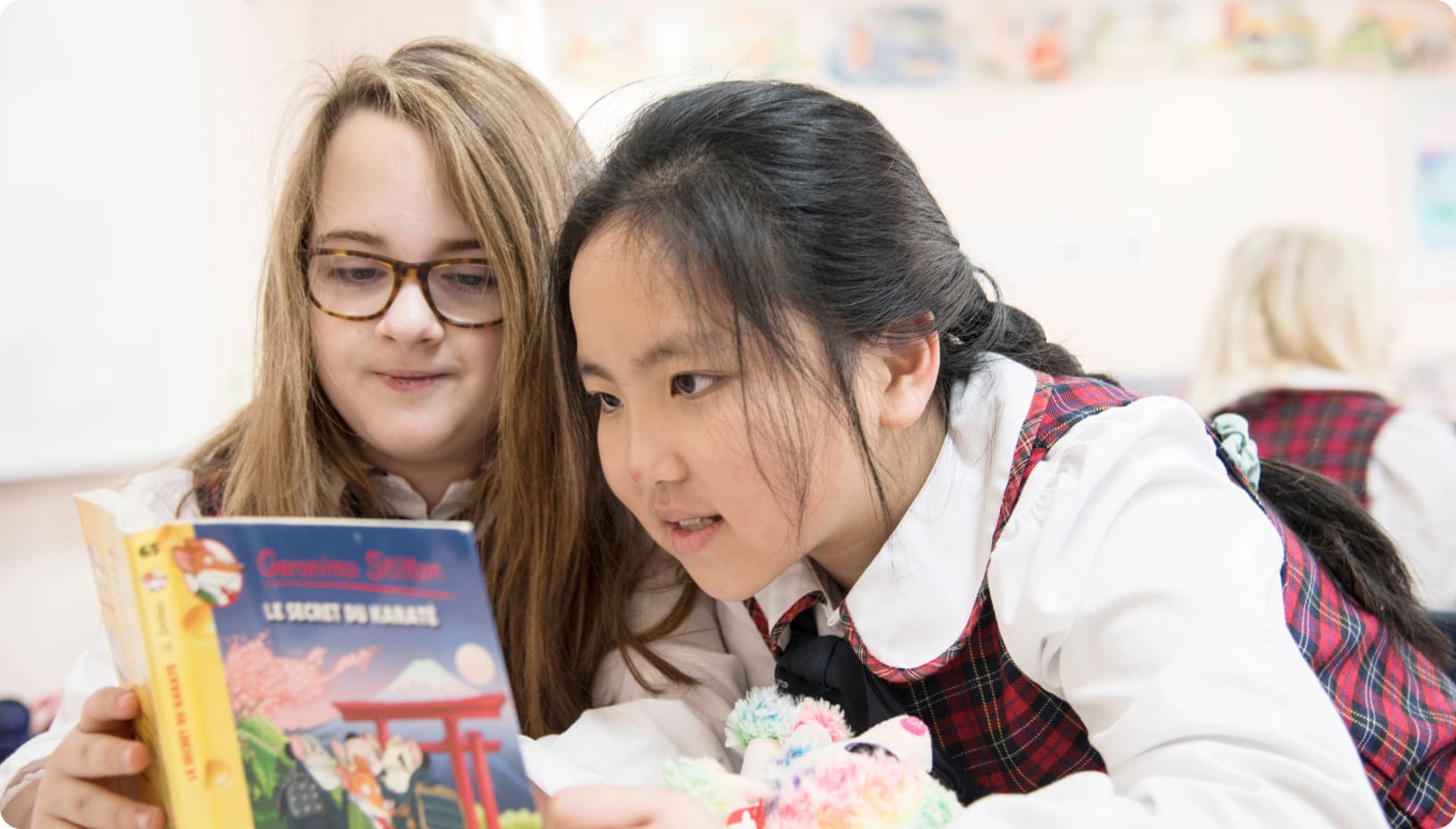 Two girls reading together, focused on a book titled "Le Secret du Sabaku." They are in a classroom, highlighting collaboration and learning.