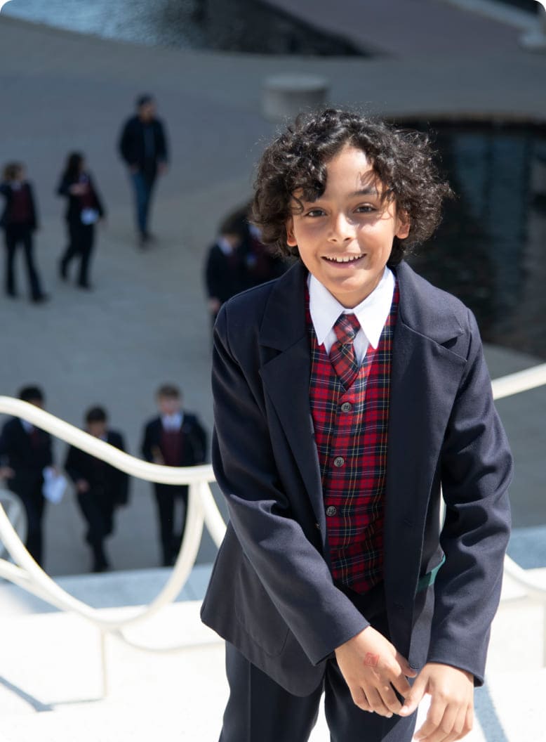 Smiling boy in a formal outfit with a plaid vest poses on steps, while other students in the background walk toward a water feature.