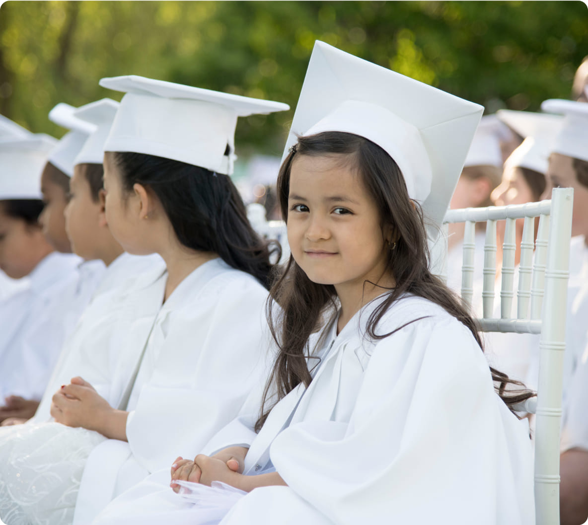 Young girl in a white graduation gown and cap smiles while seated among peers, capturing the joy of a graduation ceremony.