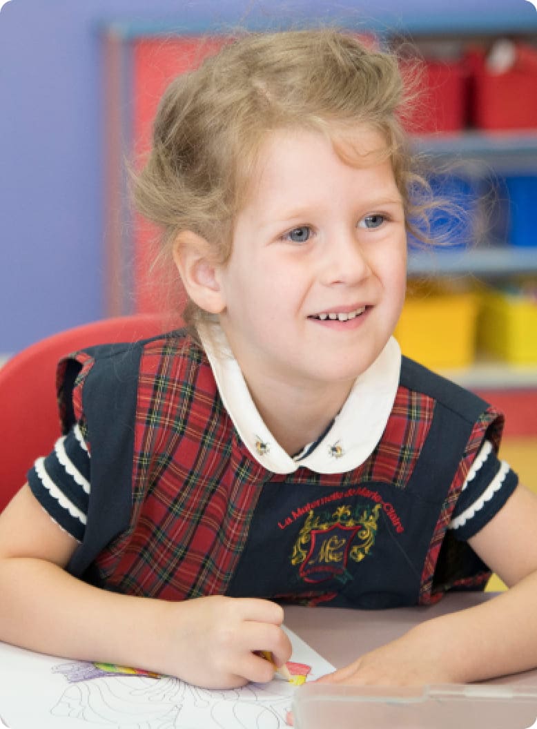 Young girl in a plaid school uniform sits at a table, smiling while coloring on a sheet of paper, reflecting a joyful classroom environment.