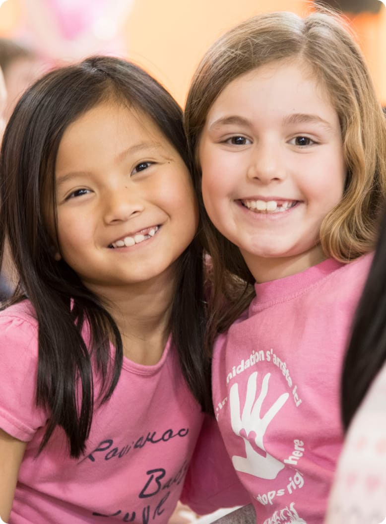 Two smiling girls wearing pink shirts pose closely together against a warm backdrop, showcasing friendship and joy in a community setting.