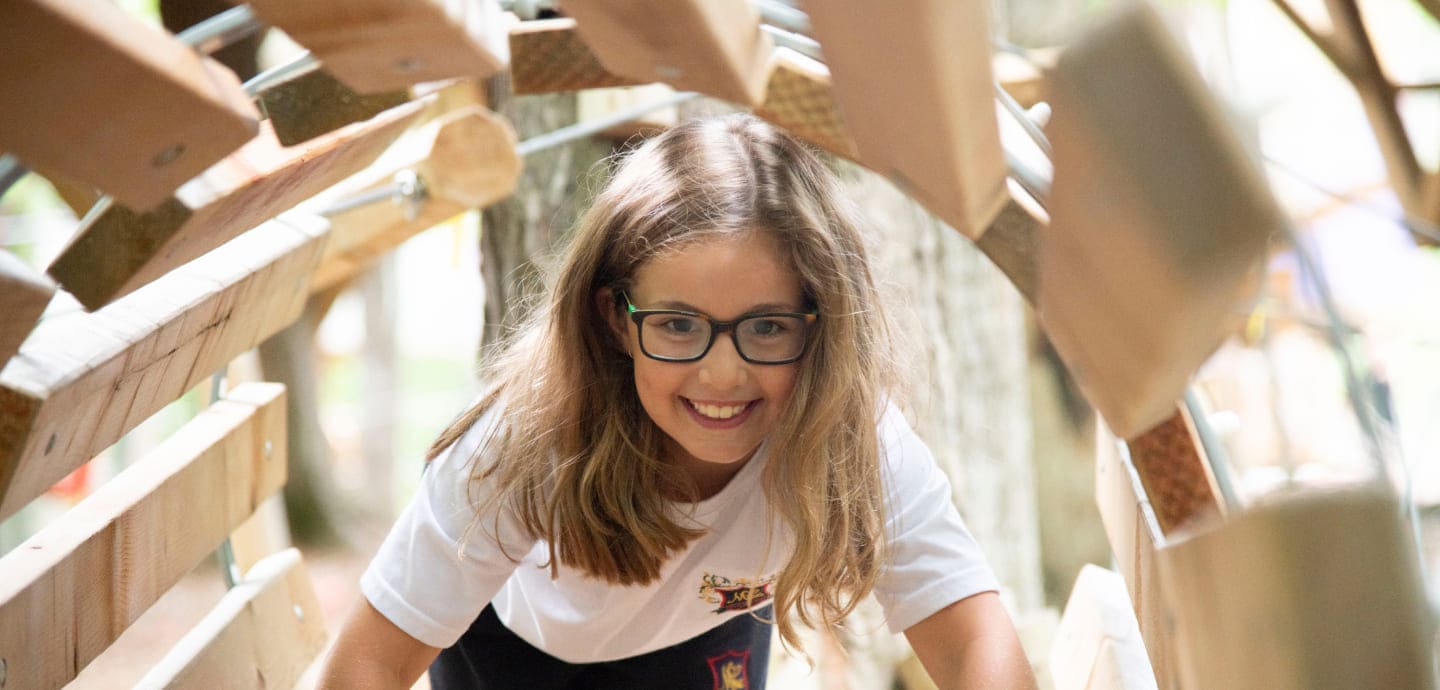 A young girl with glasses smiles while crawling through a wooden tunnel in a forest play area, showcasing joyful outdoor exploration.