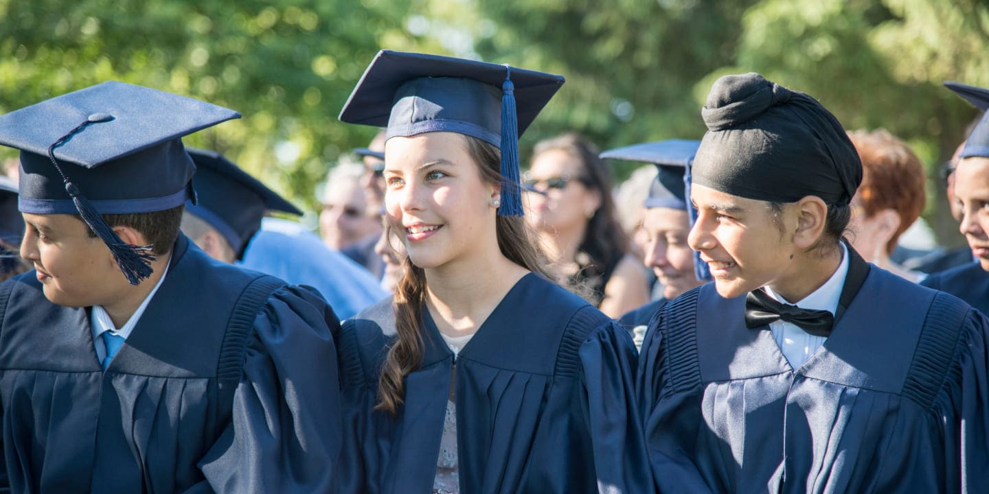 Graduates in blue caps and gowns smile during a commencement ceremony, celebrating achievement in a sunlit outdoor setting.
