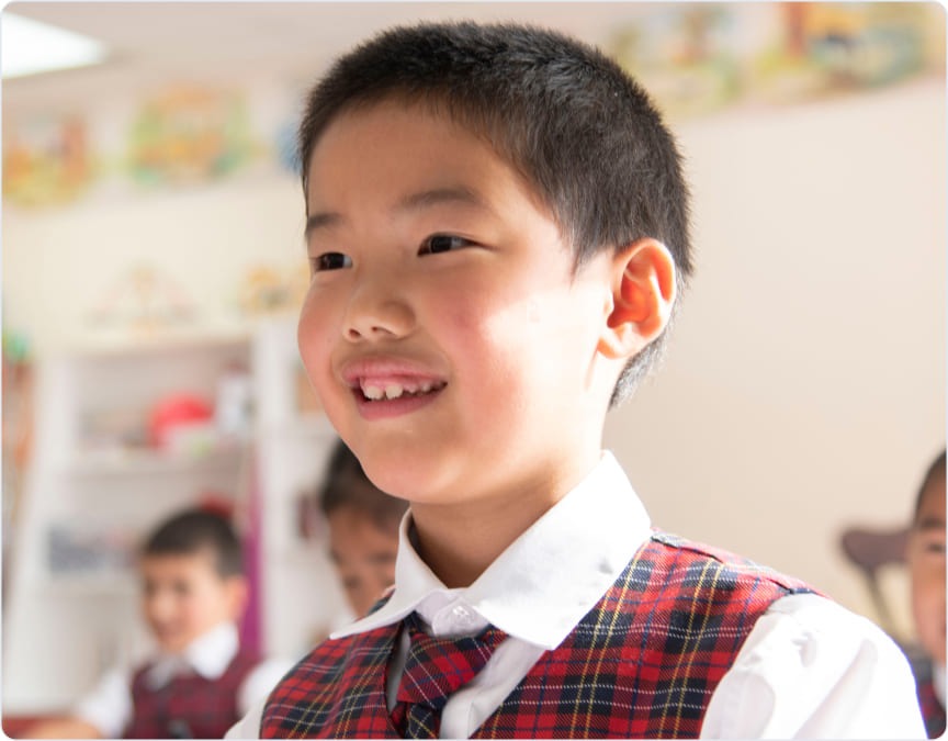 Smiling boy in a checkered vest and white shirt, engaged in a classroom activity. Colorful educational materials visible in the background.
