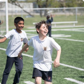 Two children run joyfully on a grassy field during a game, showcasing the excitement of outdoor play and teamwork in a school setting.