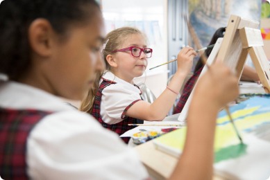 Young girl with glasses focuses on painting at an easel, while another child works in the foreground. The setting highlights a creative art class.