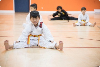 A smiling boy in a karate uniform performs a split, with peers practicing in the background. The scene highlights kids engaged in martial arts training.