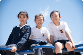 Three smiling girls in school uniforms sit on playground equipment against a clear blue sky, enjoying outdoor playtime.