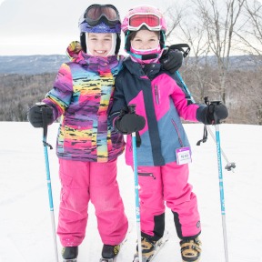 Two young girls, smiling in colorful ski outfits, stand on a snowy slope with ski poles. Their joyful expressions capture the excitement of winter sports.