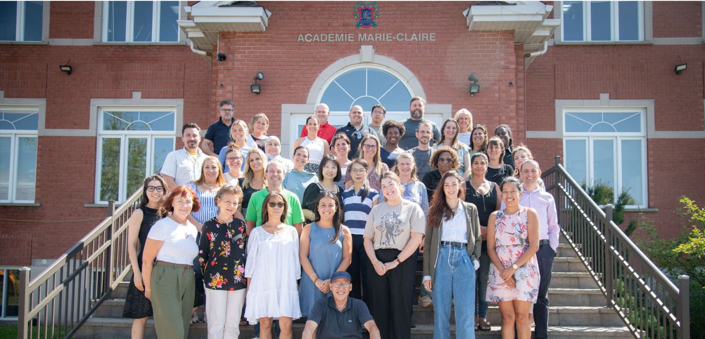 Group photo of faculty and staff from Académie Marie-Claire, standing on steps in front of the school building under clear skies.