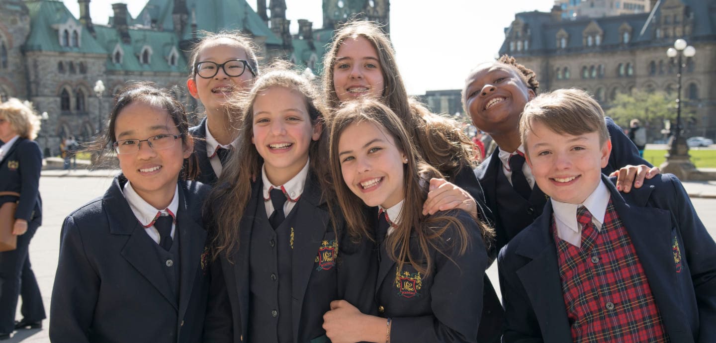 A group of seven smiling students in formal attire poses together outdoors, with historical buildings in the background, showcasing camaraderie and joy.