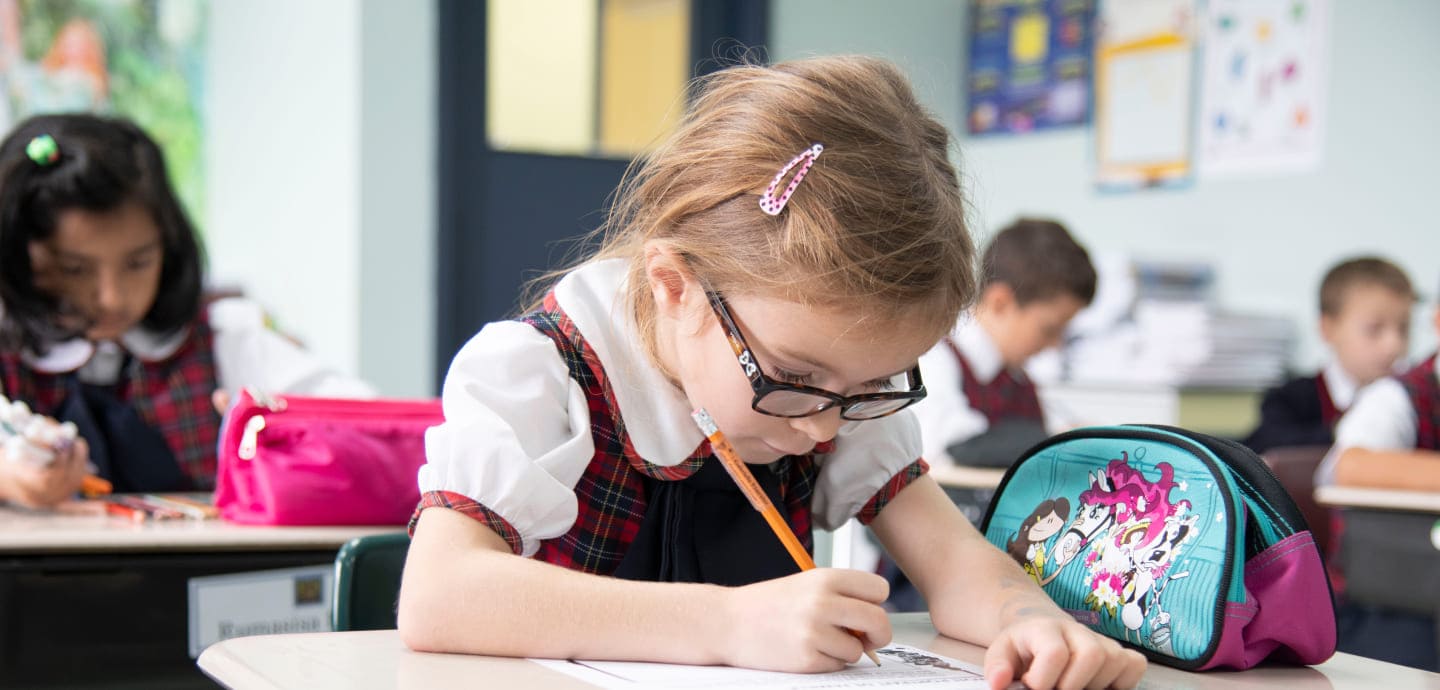 A young student with glasses concentrates on drawing in a classroom while peers work nearby. Brightly colored school supplies are visible.